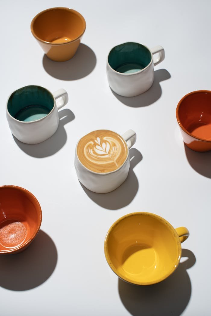 contact-img Top view of colorful ceramic cups and a latte with intricate latte art on a flat surface.